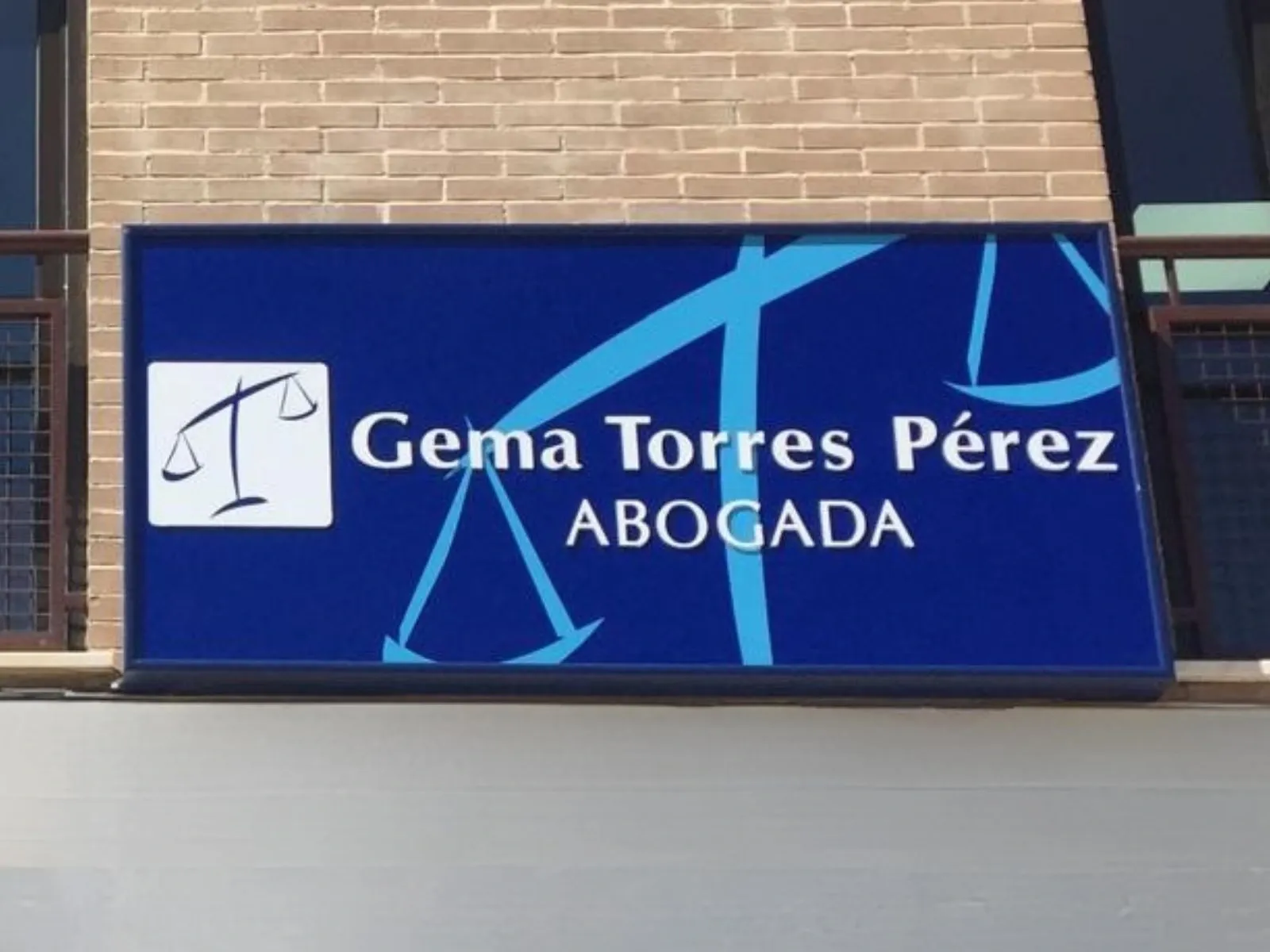 Caja de luz rectangular con frontal azul, una balanza y el nombre de la abogada Gema Torres Pérez, instalada sobre la fachada de ladrillo de un despacho